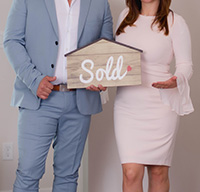 Couple dressed in nice clothes giving thumbs up holding a house-shaped Sold Sign after buying a home in the Ortley Beach real estate market